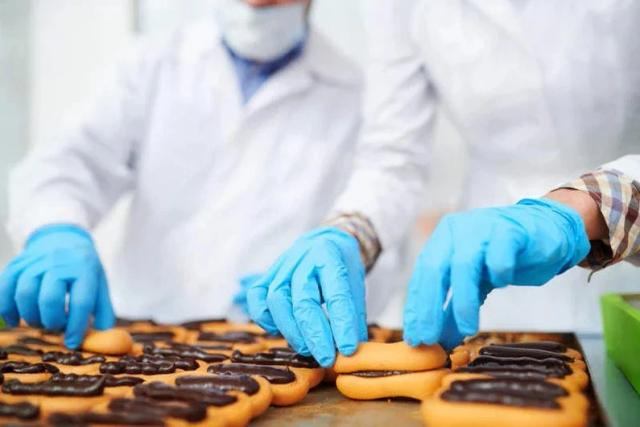Workers wearing blue nitrile gloves inspect and handle chocolate-coated pastries on a production line, illustrating industrial food processing, packaging hygiene, and potential chemical exposure risks in commercial baked goods.