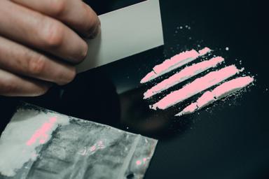 Close-up of a hand preparing lines of pink cocaine on a dark surface, with a bag of the pink substance in the foreground, highlighting themes of substance abuse, drug preparation, and public health awareness