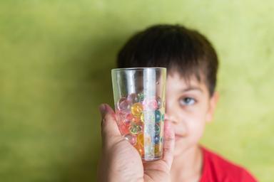 Child holding a transparent cup filled with colorful water beads against a green background, illustrating the potential hazards of water beads for young children