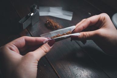 Close-up of hands preparing a substance, identified as pink cocaine, on a wooden table. The individual is delicately rolling the substance in paper under dim lighting, emphasizing the recreational and clandestine context. Ideal for articles addressing substance use, recreational drugs, or drug culture awareness