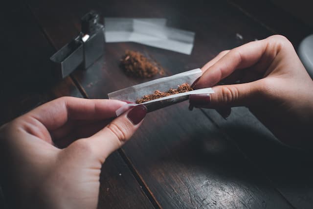Close-up of hands preparing a substance, identified as pink cocaine, on a wooden table. The individual is delicately rolling the substance in paper under dim lighting, emphasizing the recreational and clandestine context. Ideal for articles addressing substance use, recreational drugs, or drug culture awareness