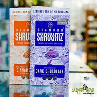 The image displays two packages of Diamond ShruumzTM brand microdosing chocolate bars. The left package is labeled "Cinnamon" and the right one is "Dark Chocolate." Both packages feature the text "Enhanced Mushrooms" and "Leading Form of Microdosing." The