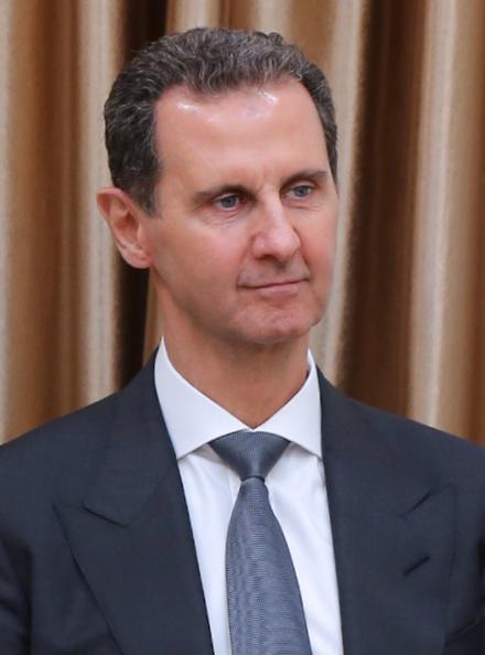 Portrait of Bashar al-Assad wearing a dark suit and tie, standing in front of beige curtains, with a composed expression and neatly combed hair